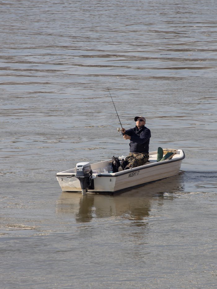 Home A man engages in fishing from a motorboat, casting his line into calm waters from a high angle view.