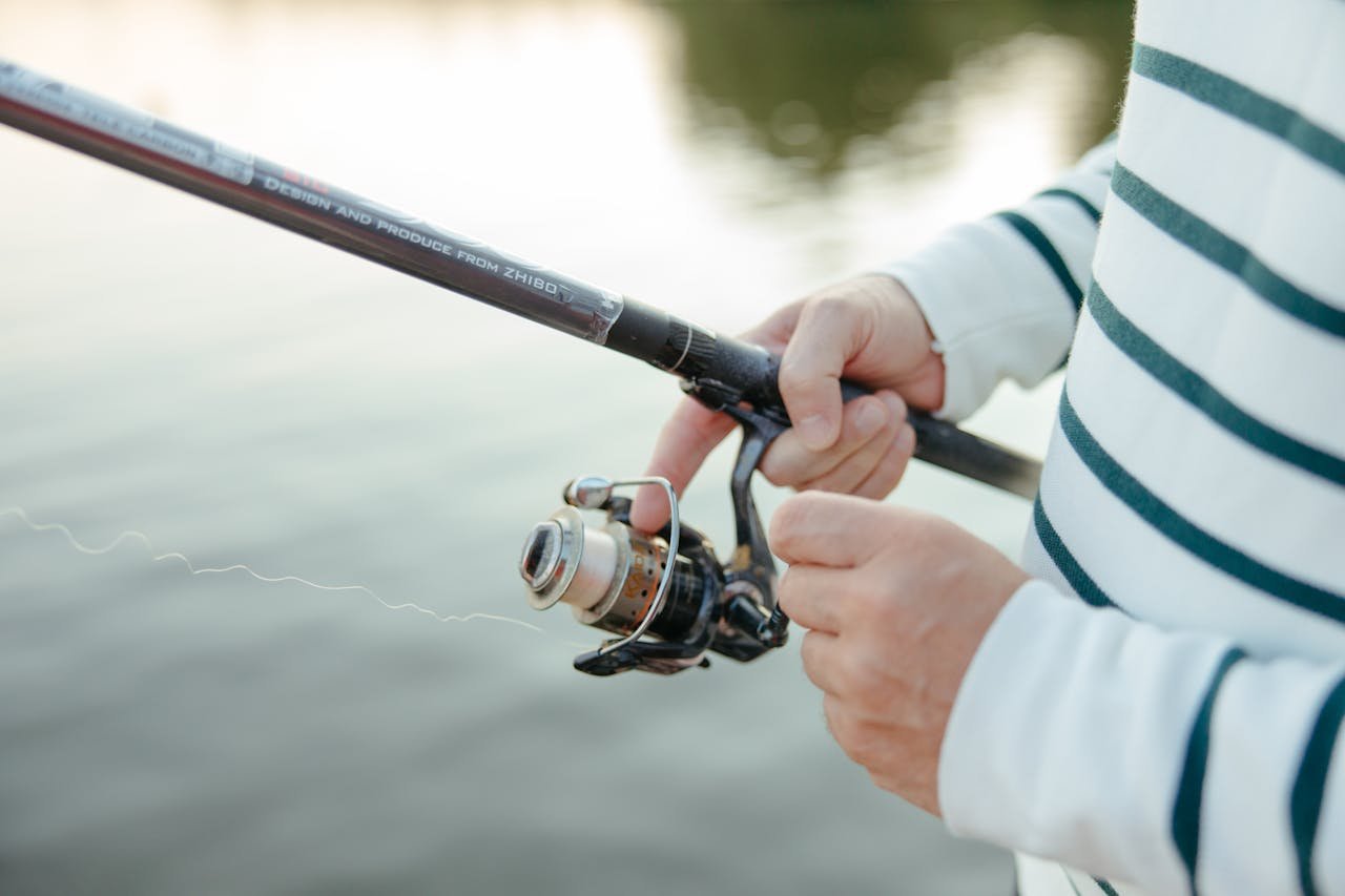 Close-up of hands holding a fishing rod with a wheel by the water, capturing leisure and recreation.